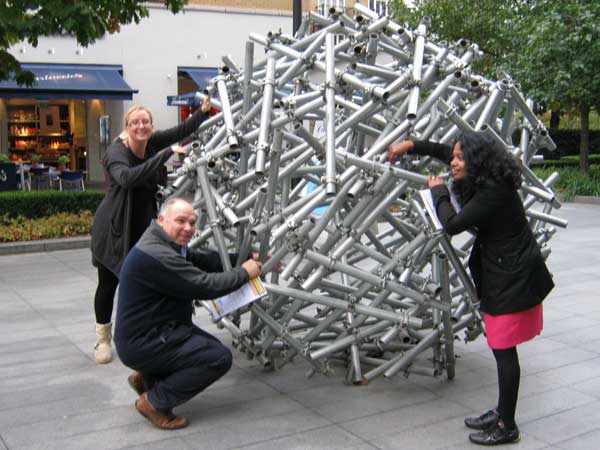 Three people search for a clue on a sculpture made of scaffolding.